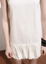 Vestido corto de algodón color marfil, sin mangas, con cuello redondo y diseño recto que presenta un bajo abullonado. Cierre de botón en la parte posterior e incluye forro interior.