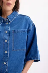 Sobrecamisa de jean de manga corta y corte oversize en color azul medio, con bolsillos frontales con solapa y cierre de botones metálicos a presión.