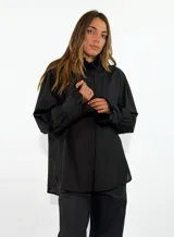 Camisa negra de corte oversize, con cuello alto con volados y mangas largas con puños elásticos también decorados con volados. Presenta cierre frontal oculto por tapeta.