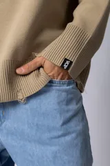 Sweater oversize de tejido de algodón color beige, con cuello redondo, puños y ruedo acanalados con detalles de desgaste. Presenta un diseño minimalista con etiqueta distintiva en el puño.