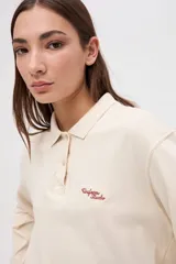 Remera tipo polo de manga larga en color beige, confeccionada en algodón pesado con textura suave. Presenta cuello clásico, cierre frontal con botones y un pequeño bordado en el pecho. Posee puños en las mangas y un corte relajado.