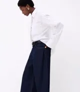 Camisa de mujer de corte maxi oversized, confeccionada en tricolina de algodón certificado. Presenta rayas verticales de hilo teñido, cuello con corbata (gravata), botones frontales, bolsillo en el pecho y puños largos.