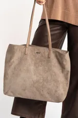 Bolso tipo tote de gran tamaño en color camel, confeccionado en cuero vegano con acabado efecto gamuza. Presenta doble asa larga para llevar al hombro y una placa metálica pequeña con el logo de la marca en el frente.