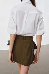 Camisa de algodón con cuello solapa y mangas por debajo del codo. Presenta un diseño con pechera plisada frontal, bajo recto y cierre mediante botones ocultos bajo solapa.