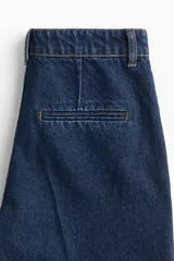 Jean tobillero de denim rígido en tono azul oscuro, con corte de tiro alto, diseño curvo y voluminoso en las piernas, y pliegues frontales y traseros.