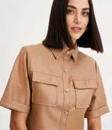 Camisa de manga corta confeccionada en cuero sintético color tostado, con cuello camisero, cierre frontal con botones en forma de corazón y dos bolsillos con solapa en el pecho.