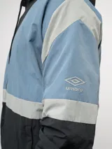 Campera de nylon Umbro con capucha, cierre frontal y diseño de bloques de color en gris, blanco y azul. Puños y dobladillo elásticos.