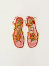 Sandalias planas de cuero color marrón, con diseño de punta cuadrada, tiras finas cruzadas y un detalle trenzado circular en el empeine. Cuentan con tiras para atar al tobillo decoradas con cuentas turquesas en las puntas.