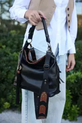 Cartera tipo hobo de cuero vacuno negro con detalles en cuero color tabaco. Presenta una correa ancha decorada con apliques metálicos, borlas laterales y herrajes en bronce envejecido. Incluye cierre principal superior y compartimentos organizadores internos.