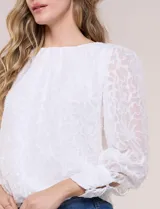 Blusa blanca de organza con textura devoré floral, silueta semiholgada y forro interno. Tiene escote bote, mangas largas abullonadas con transparencias, puños anchos con botones y ruedo elastizado.