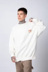 Buzo de tejido friza color crema, de corte oversize y cuello redondo. Presenta un pequeño bordado o parche en el pecho izquierdo con un diseño de cabeza de animal en tonos rojizos/terracota.