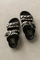Sandalias de cuero de doble tira, con estampa animal print tipo vaca en tonos blanco, negro y marrón, adornadas con tachas metálicas en la tira superior y una hebilla grande en la tira inferior.