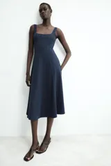 Vestido midi de color beige con escote cuadrado y tirantes anchos. Presenta un diseño entallado en el torso con costuras verticales marcadas y falda con caída evasé.
