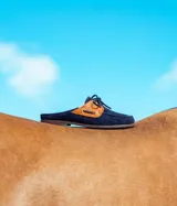 Mules estilo mocasín confeccionados en gamuza azul marino con detalles en gamuza color suela en los laterales. Presentan cordones frontales decorativos y una silueta abierta en el talón.