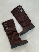 Botas de caña media en color marrón chocolate, confeccionadas en material tipo gamuza. Presentan un diseño holgado con efecto arrugado y tres correas decorativas horizontales en la parte superior con hebillas metálicas plateadas. Cuentan con suela plana y tacón bajo de goma negra.