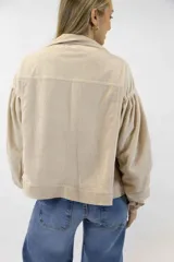 Campera corta de pana color beige, con cuello clásico, cierre frontal con botones y amplios bolsillos con solapa en el pecho. Cuenta con mangas largas abullonadas y diseño holgado.