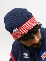 Gorro tipo beanie de punto acanalado en color azul marino, con vuelta en color rojo y blanco a rayas. Incluye etiqueta lateral con el logo de Umbro y el escudo del Club Nacional de Football.