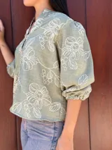 Camisa de corte holgado en color verde con estampado floral lineal en tono crema. Presenta cuello mao, cierre frontal con botones y mangas tres cuartos con puños elásticos.