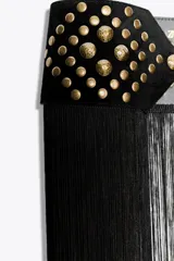 Cinturón tipo fajín negro de 10 cm de ancho, confeccionado en piel 100%. Presenta una sección superior con tachas doradas y ojales metálicos, y la parte inferior terminada en flecos largos. Se ajusta al frente con una lazada de cordón negro.
