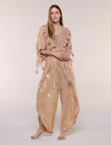 Pantalón palazzo confeccionado en gasa transparente color rosa con diseño estampado metalizado dorado. Presenta cintura elástica con cordón ajustable con borlas, piernas anchas y diseño cruzado al frente con aberturas laterales y detalles de puntillas.