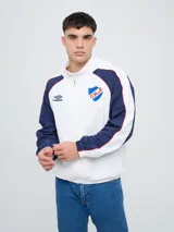 Campera deportiva Umbro del Club Nacional de Football, modelo Venti, para hombre. Diseño en color blanco con mangas azules y detalles de líneas rojas. Cuenta con capucha con cordón ajustable, cierre frontal y bolsillos. Presenta el logo de Umbro y el escudo del club bordados en el pecho.