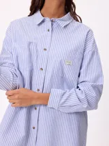 Camisa de corte oversize con diseño a rayas verticales en tonos azul y blanco. Presenta cuello clásico, cierre frontal con botones y un bolsillo en el pecho con detalle de etiqueta.