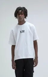 Remera blanca de corte oversize y hombros caídos, confeccionada en algodón premium. Presenta cuello redondo acanalado y estampado frontal con las siglas KM y el hashtag #KILOMBITOMAL en color negro.