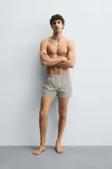 Bañador corto de hombre con cintura elástica ajustable mediante cordón blanco. Presenta un estampado geométrico repetitivo en tonos oscuros (negro/marrón oscuro) y blanco/beige. Incluye bolsillos laterales y un bolsillo trasero de vivo.