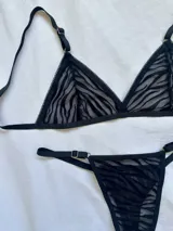 Conjunto de lencería de dos piezas confeccionado en tul transparente con estampado animal print tipo cebra aterciopelado en color negro. Incluye un corpiño triangular sin aro con breteles regulables y una bombacha tipo tanga a juego con detalles de argollas metálicas en los laterales.