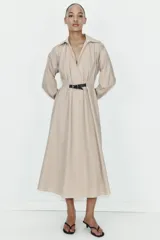Vestido midi de color beige con cuello solapa y escote en pico. Presenta mangas largas, cintura elástica en la espalda y un cinturón a contraste con hebilla metálica. Incluye bolsillos laterales ocultos.