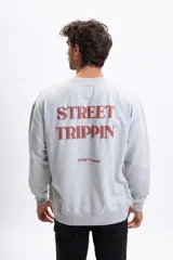 Buzo de cuello redondo color gris melange, con estampa frontal en color bordó con la frase 'STREET TRIPPIN' y logo 'STRP Capsule'. Presenta un diseño de corte holgado con puños y ruedo acanalados.