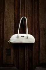 Cartera tipo baguette color blanco, confeccionada en cuero, con manijas largas y detalles de nudos esféricos de alpaca en la unión de las manijas. Presenta una silueta alargada y compacta.