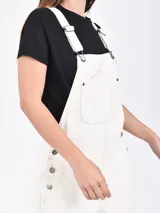 Monoprenda corta estilo overol de jean color beige, con bolsillos frontales, tirantes ajustables con hebillas metálicas y short con vuelta en el dobladillo.