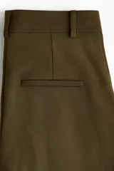 Pantalón verde militar de corte holgado, con pinzas delanteras, bolsillos laterales, bolsillos traseros decorativos y piernas amplias con pliegues marcados. Cuenta con cierre de botón y corchetes ocultos.
