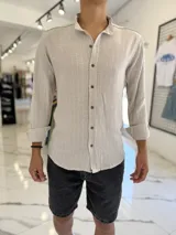 Camisa de lino color tostado con sutiles rayas verticales blancas, cuello mao y mangas largas con puños vueltos. Cuenta con cierre frontal de botones oscuros.