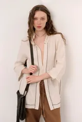 Chaqueta beige de lino con mangas 3/4, cuello mao y detalles de costuras decorativas oscuras en contraste.