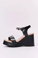Sandalias de cuero negras con plataforma y tacón de 10 cm. Tienen una tira ancha en la parte delantera y una tira ajustable con hebilla alrededor del tobillo, ambas con detalles de tachas plateadas.