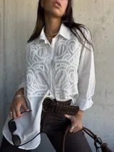 Camisa blanca de corte oversize con diseño de bordado calado en el frente. Presenta cuello clásico, cierre frontal con botones y mangas largas.