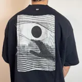 Remera negra de corte oversize con estampa rectangular en la espalda que representa una mano alcanzando un círculo sobre un fondo de líneas horizontales.