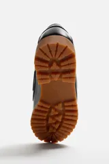 Zapato tipo náutico de cuero color marrón, con cordones, ojales metálicos laterales y suela de goma con volumen y relieve dentado.