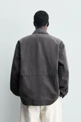 Cazadora relaxed fit de hombre, confeccionada en tejido técnico con algodón de color negro. Cuenta con cuello camisero, manga larga y bolsillos frontales de plastrón con solapa. Presenta cintura ajustable con cordón interior y cierre frontal con cremallera oculta por solapa con botones a presión.