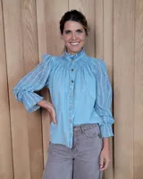 Camisa de denim celeste con detalles de broderie en las mangas, cuello alto con volados y canesú con frunces. Presenta cierre frontal con botones y mangas largas con puños elásticos.