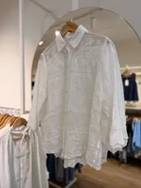 Camisa blanca de calce oversize, con cuello clásico y cierre frontal de botones. Presenta delicados bordados en relieve distribuidos por toda la prenda, mangas largas y ruedo asimétrico.