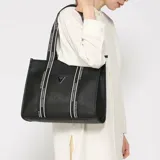 Cartera tipo tote Guess modelo Jackie, de color negro con textura saffiano y asas de cinta estampada con texto "Guess Los Angeles". Cuenta con un bolsillo frontal exterior y aplique metálico triangular con el logo de la marca.