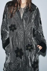 Kimono de terciopelo gris con acabado texturizado, cuello redondo y manga larga. Presenta bordados florales en hilo negro y metalizado, bolsillos delanteros y cierre frontal.