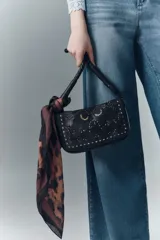 Cartera de mano tipo baguette confeccionada en cuero vacuno negro, con solapa decorada mediante diseño calado y tachas metálicas. Presenta una correa trenzada y un pañuelo estampado anudado en uno de sus laterales.