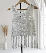 Chaleco sin mangas de crochet blanco, tejido con un patrón de círculos o flores en relieve. Presenta un borde inferior con flecos largos.