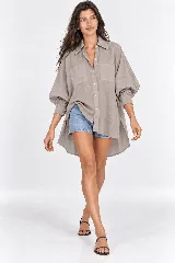 Camisa oversize color gris topo, confeccionada en 100% lino, de corte asimétrico con espalda más larga. Presenta cuello clásico, cierre frontal con botones, bolsillos plaque en el pecho y mangas abullonadas con puños estructurados con botones y pliegues.