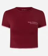 Remera deportiva cropped de manga corta, color rojo oscuro, con cuello redondo y ribetes contrastantes en blanco en el cuello y los puños. Presenta una pequeña inscripción en el pecho.