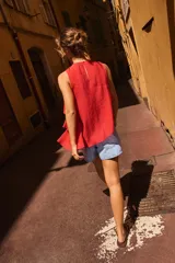 Top de cuello redondo y manga sisa, confeccionado en tejido con acabado satinado. Presenta volantes en cascada y un diseño de bajo asimétrico, con cierre en la espalda mediante pequeña abertura y botón.